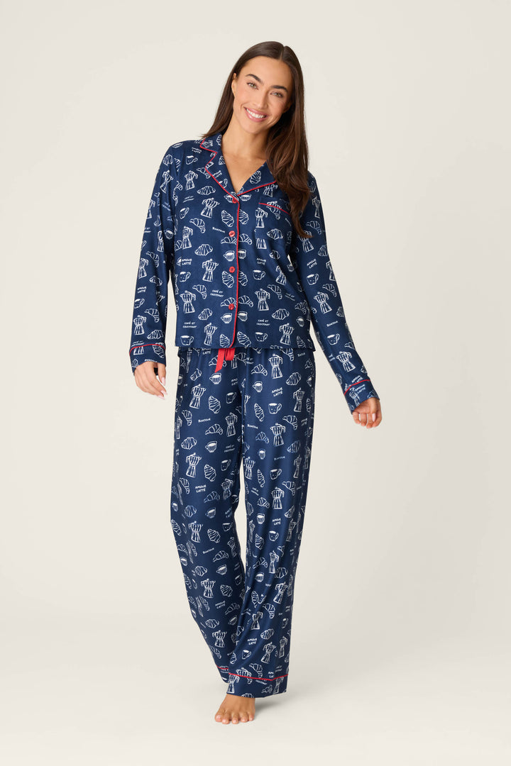 A woman stands barefoot and smiling in a P.J. Salvage Bonjour Babe French Breakfast Print Long Sleeve PJ Set, featuring a matching navy blue top with white and red patterns, button closure, long sleeves, and loose-fitting pants. The background is plain and light-colored.
