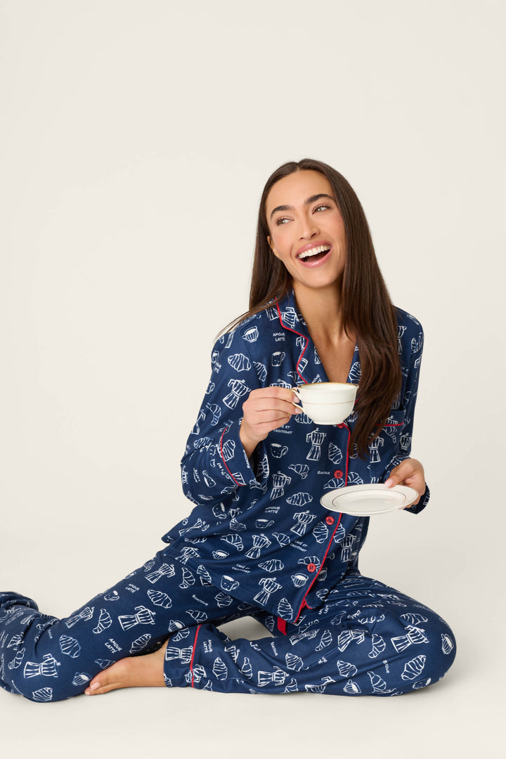A smiling woman sits on the floor in the P.J. Salvage Bonjour Babe French Breakfast Print Long Sleeve PJ Set, holding a teacup and saucer and looking off to the side against a plain light background.
