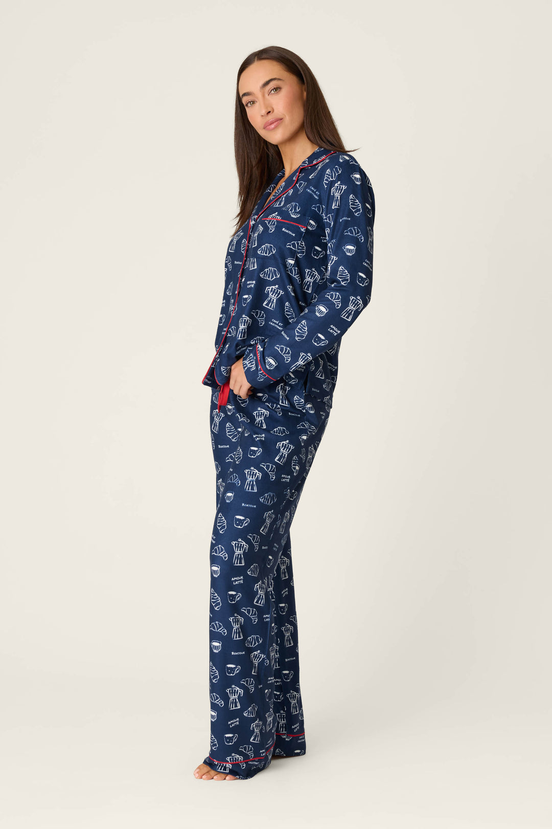 A woman stands barefoot in the P.J. Salvage Bonjour Babe French Breakfast Print Long Sleeve PJ Set, featuring a blue pajama design with playful white chef hat and utensil patterns, accented by red trim and handy pockets. She faces slightly to the side against a plain off-white background.