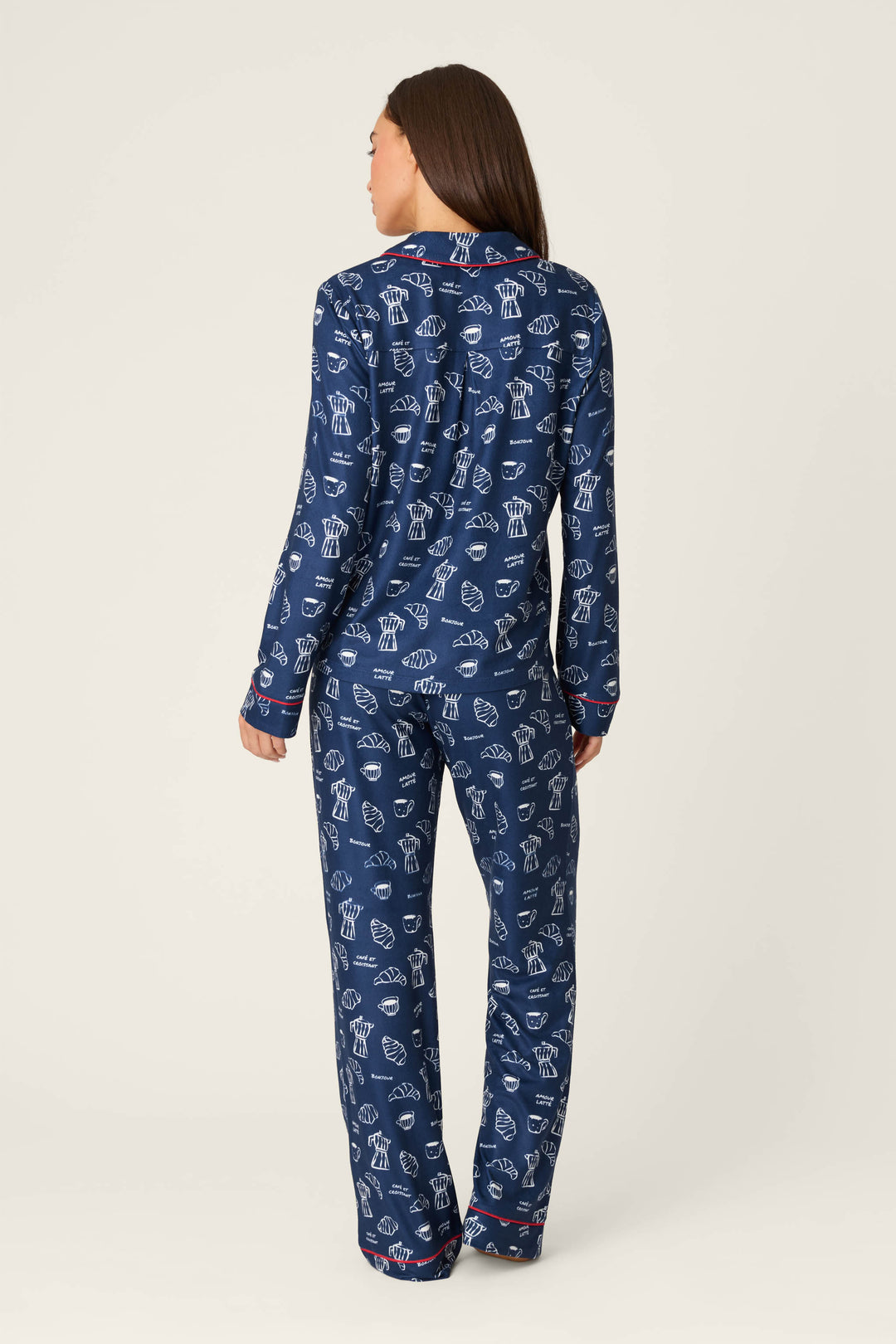 A person stands facing away, wearing the P.J. Salvage Bonjour Babe French Breakfast Print Long Sleeve PJ Set. The matching pajamas feature a navy blue background with white cupcake patterns, and include a long-sleeve top and full-length pants. The background is plain and light-colored.