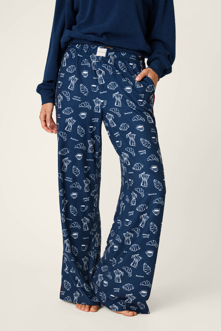 A person wearing P.J. Salvage Bonjour Babe French Breakfast Print Pajama Pants, featuring a white cupcake design on navy blue, paired with a matching navy top. The individual stands barefoot with one hand in their pocket.