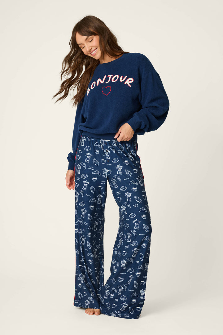 A smiling woman stands against a plain light background with one hand in her pocket, wearing a blue sweater and P.J. Salvage Bonjour Babe French Breakfast Print Pajama Pants featuring a white print.