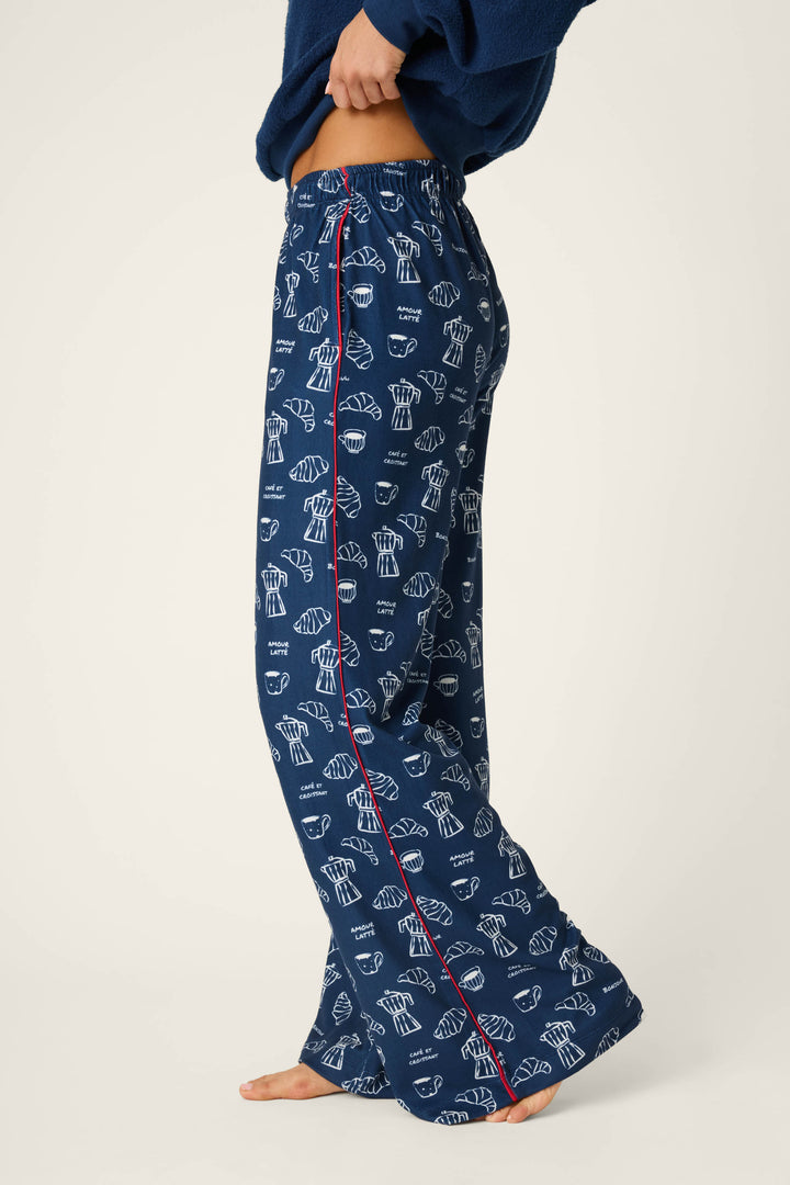A person is shown from the waist down, barefoot against a plain backdrop, wearing the P.J. Salvage Bonjour Babe French Breakfast Print Pajama Pants. The pajama pants are navy blue with whimsical white pastry and coffee cup prints and feature a red side seam; they are paired with a navy top.