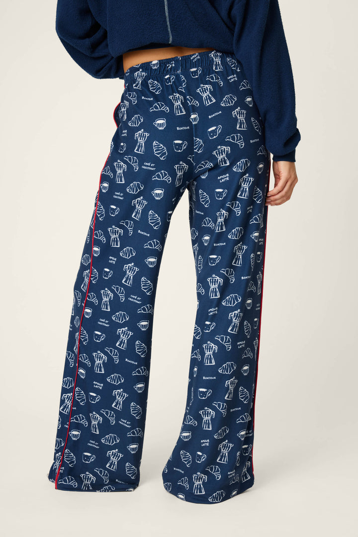 A person is wearing the P.J. Salvage Bonjour Babe French Breakfast Print Pajama Pants, featuring a navy blue base with a white croissant and bread pattern, red side stripes, shown from the back waist down and paired with a matching navy sweatshirt.