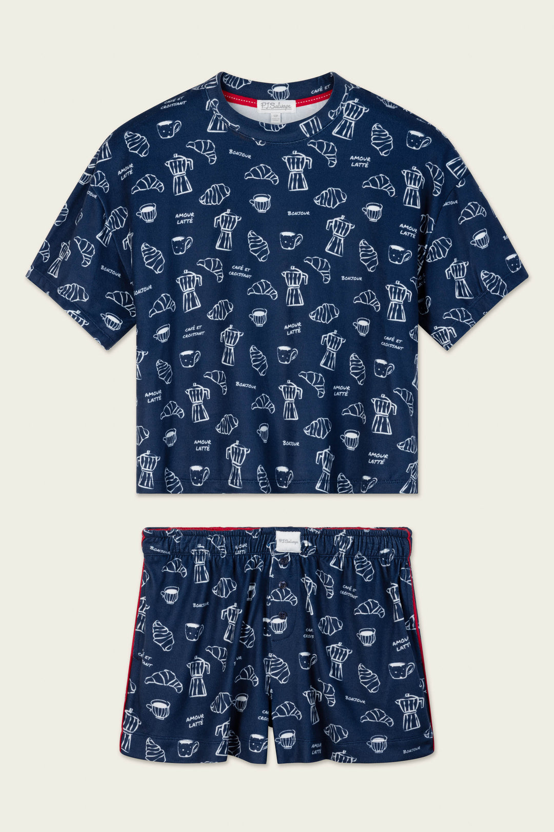 The Bonjour Babe French Breakfast Print Short PJ Set by P.J. Salvage is a navy blue pajama set with a T-shirt and shorts, featuring a playful repeating white pattern of croissants, coffee cups, and the word "Bonjour.