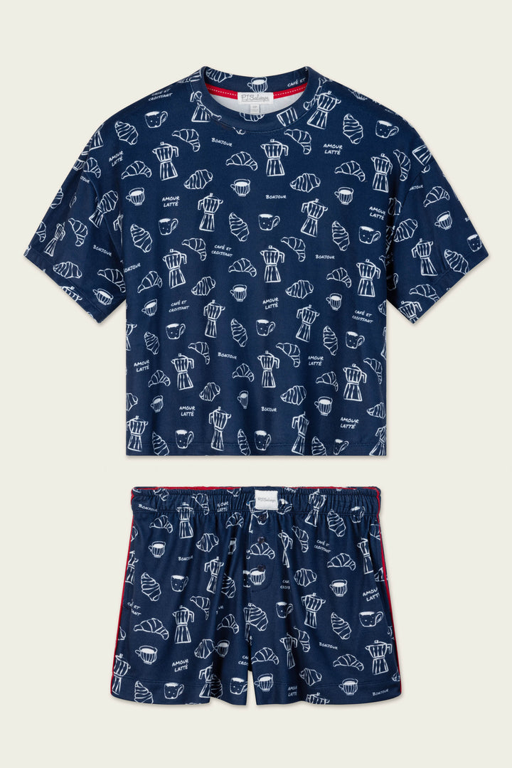 The Bonjour Babe French Breakfast Print Short PJ Set by P.J. Salvage is a navy blue pajama set with a T-shirt and shorts, featuring a playful repeating white pattern of croissants, coffee cups, and the word "Bonjour.