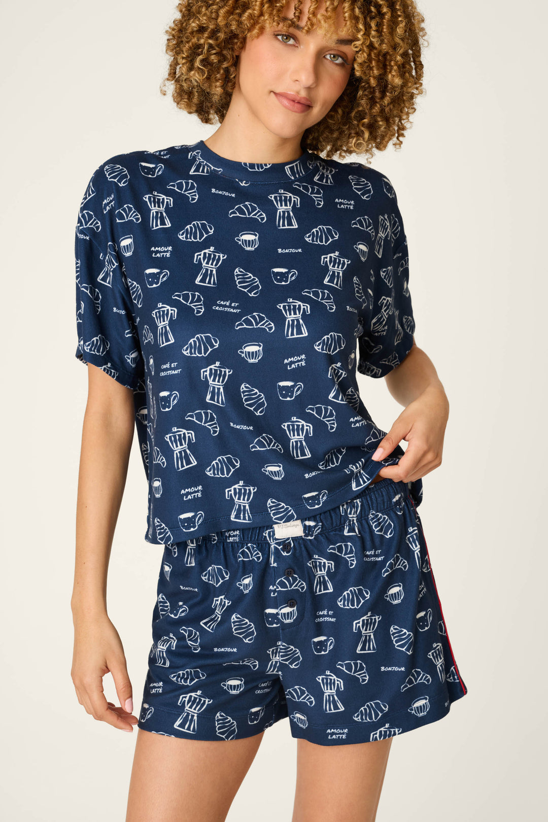 A woman with curly hair wears the P.J. Salvage Bonjour Babe French Breakfast Print Short PJ Set, featuring a matching navy blue pajama set adorned with white croissant and coffee cup patterns, as she stands against a light background holding the hem of her shirt.