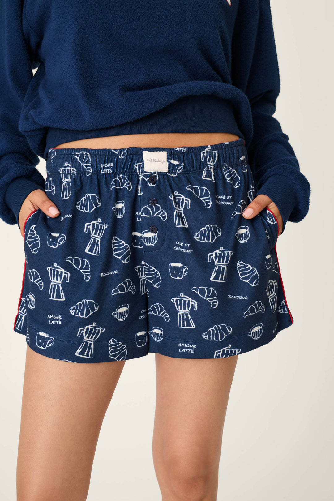 A person wears the P.J. Salvage Bonjour Babe French Breakfast Print Pajama Shorts, featuring a navy blue fabric with white line drawings of croissants, cups, coffee, and phrases like “bonjour” and “café.” The person’s hands are in the shorts’ pockets; the top half of their face is not visible.