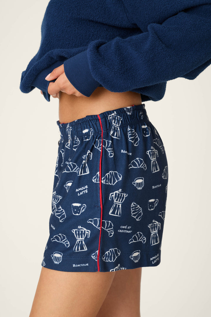 A person is shown wearing the P.J. Salvage "Bonjour Babe French Breakfast Print Pajama Shorts," which feature a blue background with white coffee and croissant illustrations, a red vertical seam, and an elastic waistband. The shorts are paired with a blue fleece top; only the torso and upper legs are visible.