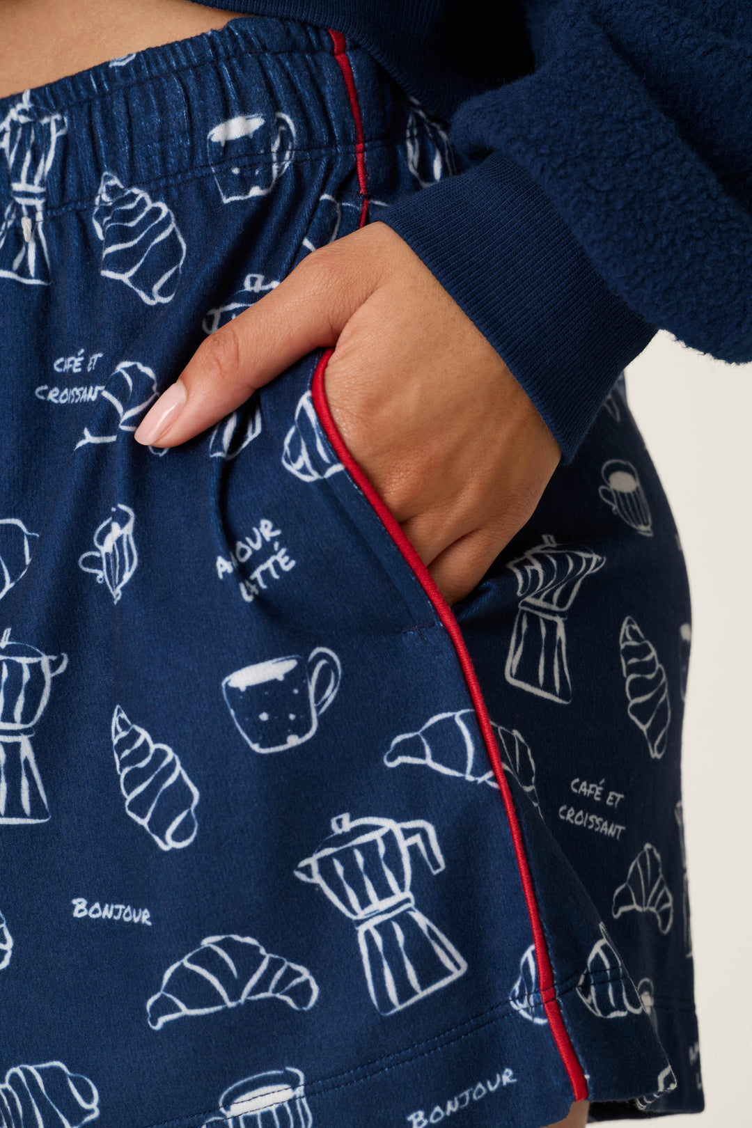 Wearing the Bonjour Babe French Breakfast Print Pajama Shorts by P.J. Salvage, featuring a navy blue fabric adorned with white croissants, coffee cups, and French phrases, this person has their hand in the pocket accented with a visible red trim.