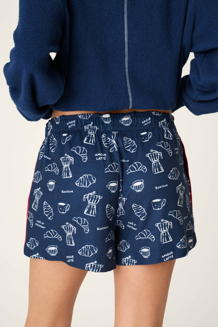 A person wearing the P.J. Salvage Bonjour Babe French Breakfast Print Pajama Shorts, featuring a navy blue fabric with white line drawings of croissants, coffee cups, and baguettes, paired with a fuzzy blue top. The image highlights the shorts from the back.