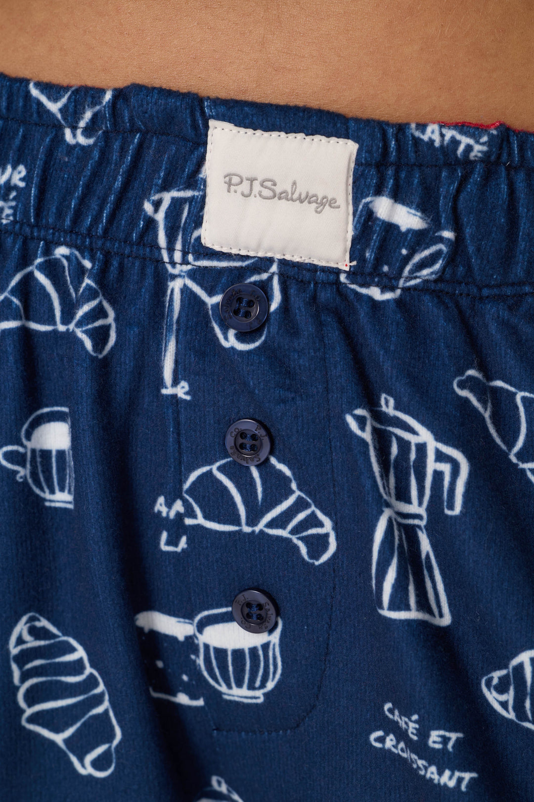 Close-up of the Bonjour Babe French Breakfast Print Pajama Shorts by P.J. Salvage, featuring a blue fabric with a white coffee and croissant pattern, three navy blue buttons, and an elastic waistband with a P.J. Salvage label.