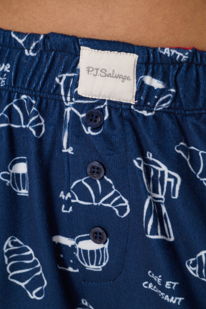 Close-up of the Bonjour Babe French Breakfast Print Pajama Shorts by P.J. Salvage, featuring a blue fabric with a white coffee and croissant pattern, three navy blue buttons, and an elastic waistband with a P.J. Salvage label.