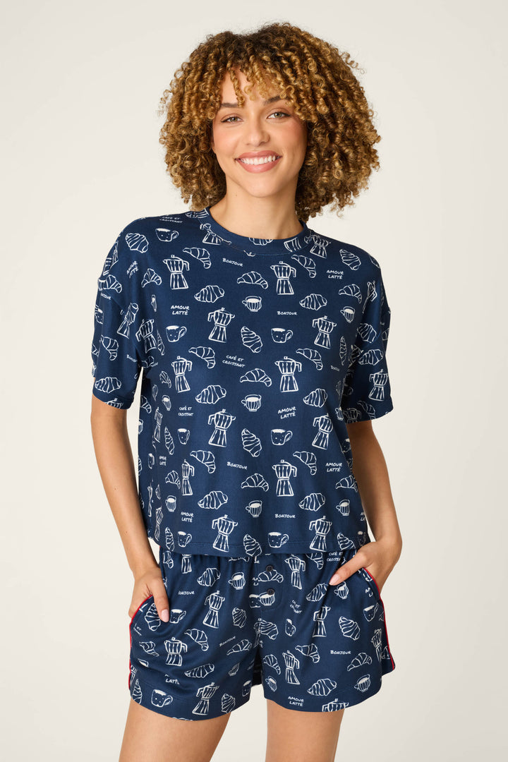 A smiling person with curly hair wears the P.J. Salvage Bonjour Babe French Breakfast Print Short Sleeve Top, paired with matching shorts featuring a white seashell pattern, hands in pockets, and stands against a plain light background.