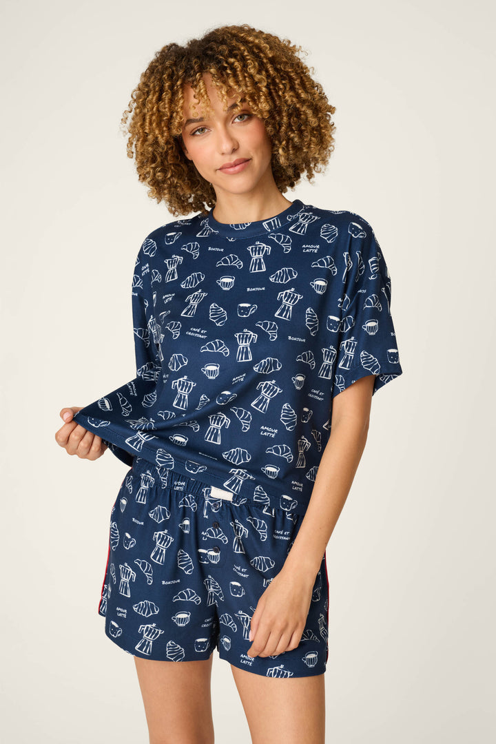 A person wearing the P.J. Salvage Bonjour Babe French Breakfast Print Short Sleeve Top, featuring a blue design with a white pastry print, holds out the hem of the shirt and smiles at the camera.