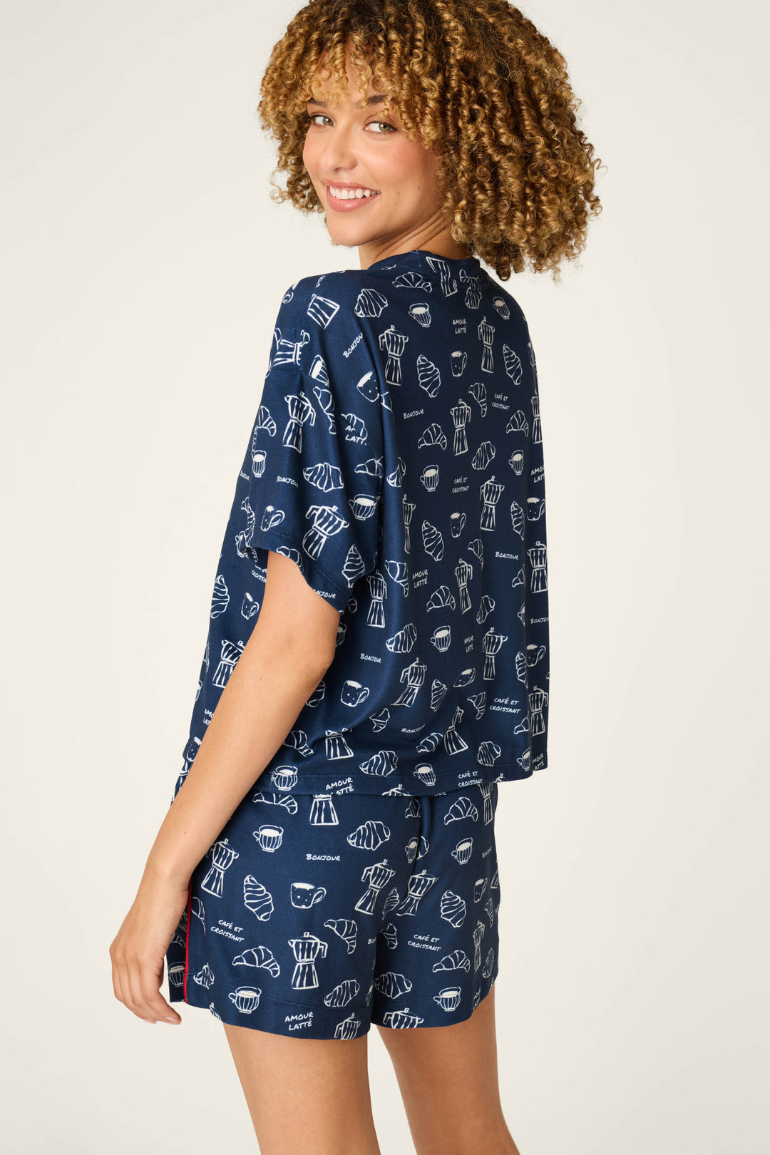 A woman with curly hair smiles as she stands sideways and looks over her shoulder against a plain background, wearing the P.J. Salvage Bonjour Babe French Breakfast Print Short Sleeve Top featuring playful white pastry illustrations on navy blue.