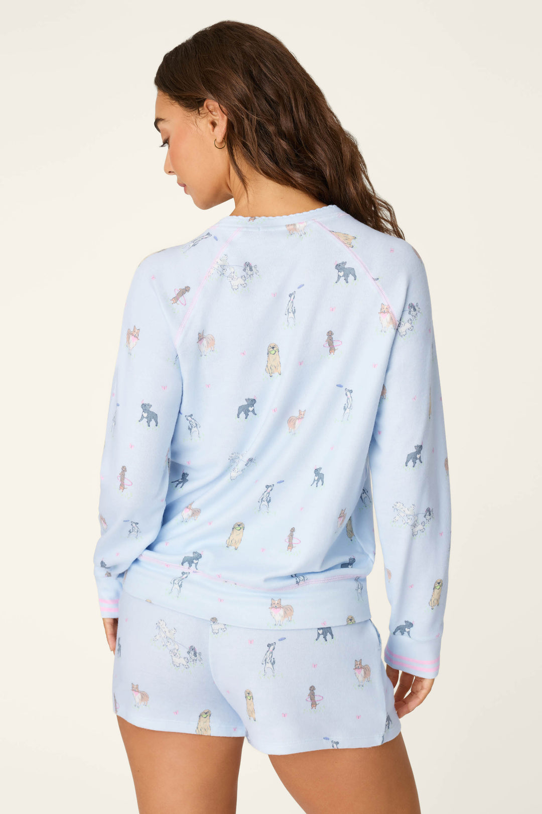 A woman with long brown hair models the P.J. Salvage Bark Park Fetch & Frolic Long Sleeve Top, a light blue, dog-print jersey pajama top, as she stands facing away and glances over her shoulder against a plain background.