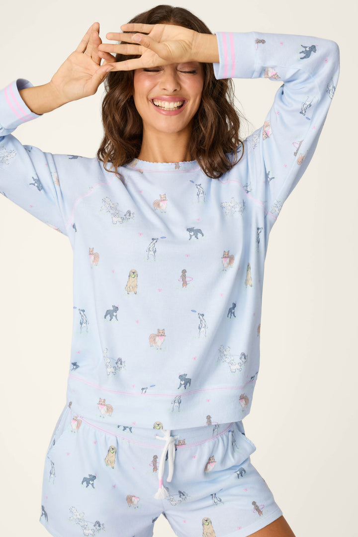 A smiling woman in the P.J. Salvage Bark Park Fetch & Frolic Long Sleeve Top playfully covers her eyes with one hand and raises the other, showing off the dog print pajama top with pink and white striped cuffs.