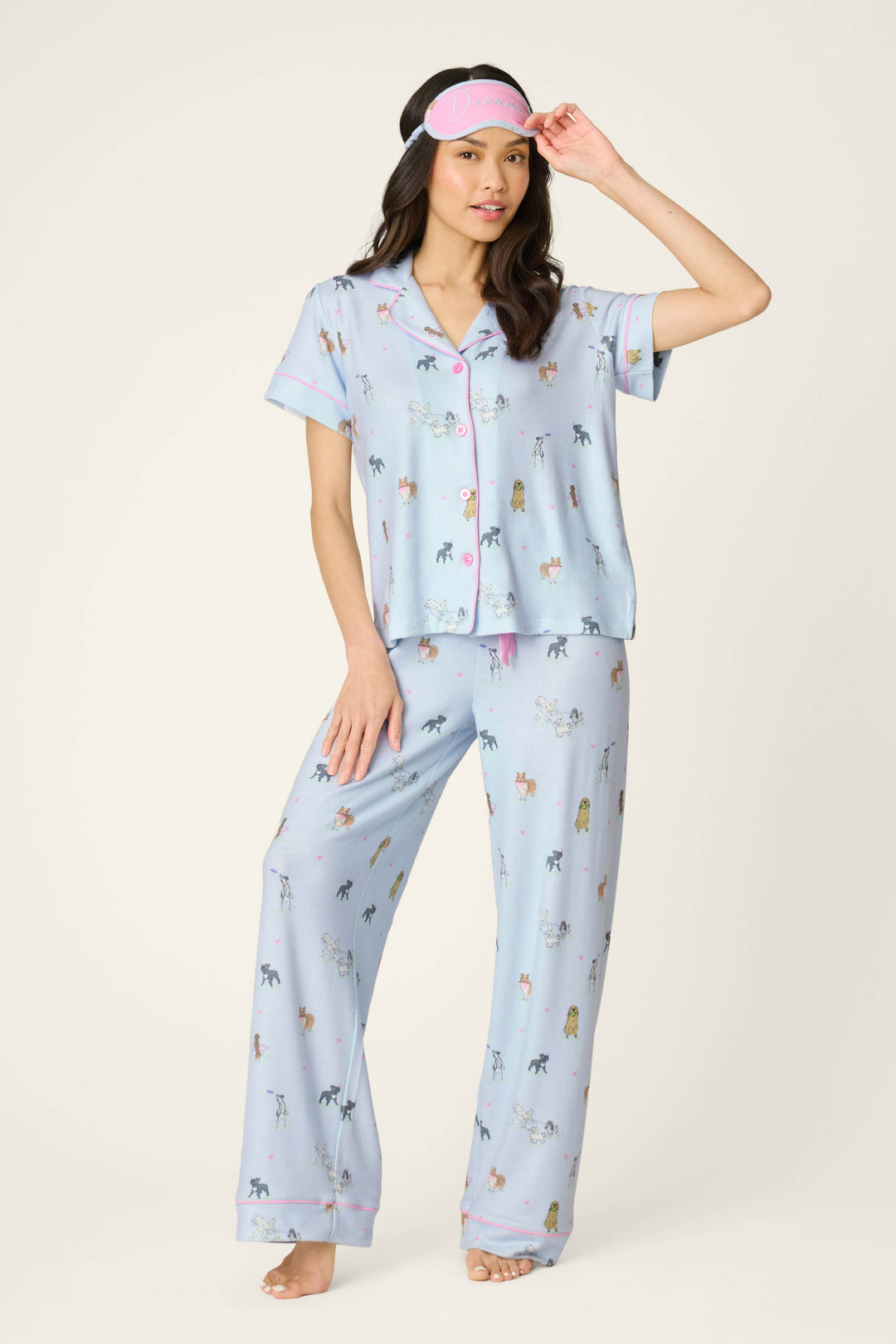 A woman models the P.J. Salvage Bark Park Fetch & Frolic 3-Piece PJ Set—light blue dog-print pajamas with matching pants and a pink sleep mask—standing barefoot, perfect for cozy women's sleepwear.