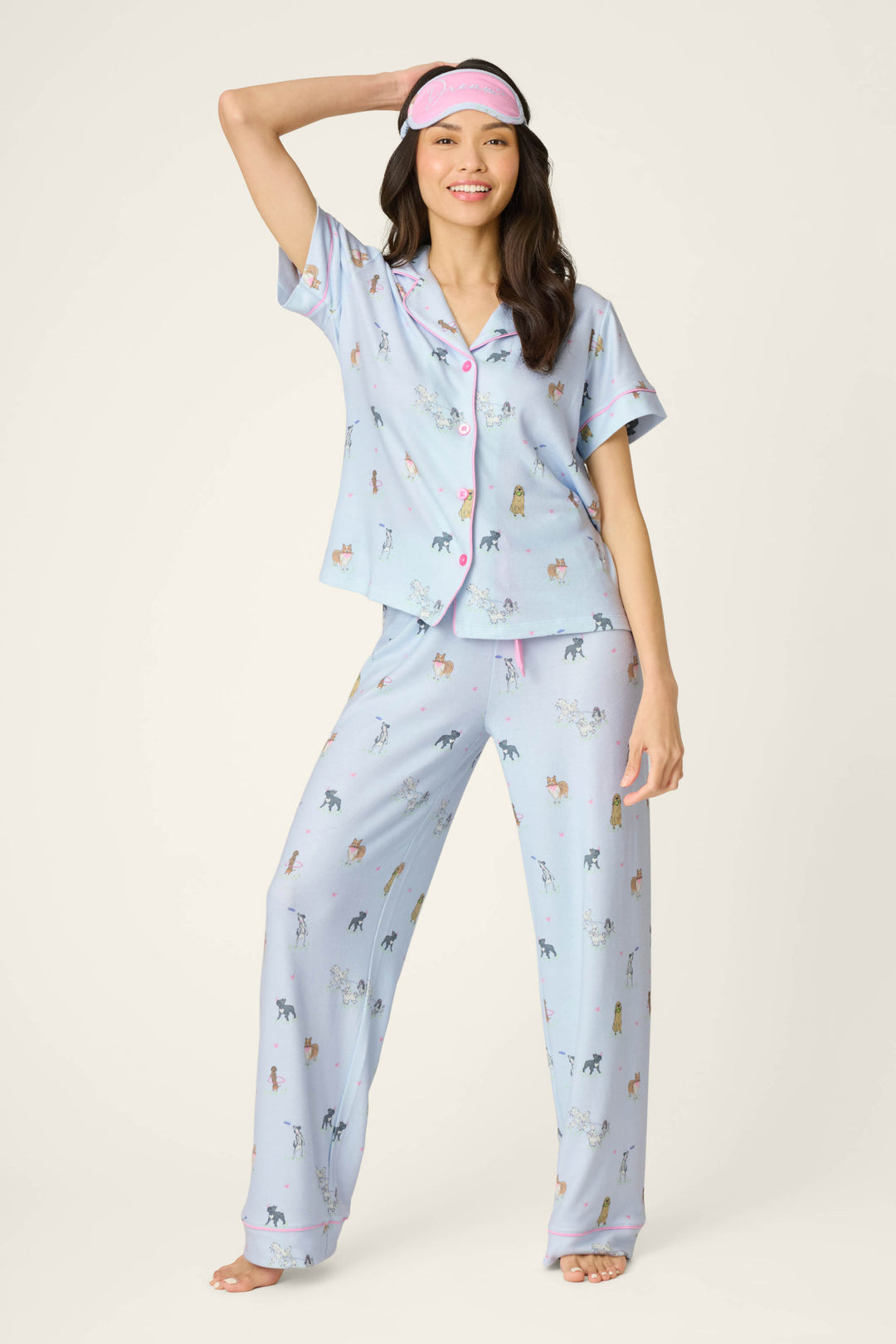 A woman barefoot and smiling stands against a plain background, wearing the P.J. Salvage Bark Park Fetch & Frolic 3-Piece PJ Set—colorful dog lover pajamas with pink trim—while holding a pink sleep mask on her head.