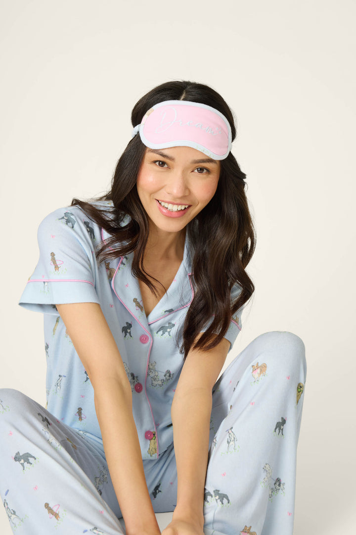 A smiling woman in the P.J. Salvage Bark Park Fetch & Frolic 3-Piece PJ Set—light blue dog lover pajamas with pink piping and a matching “Dream” sleep mask—sits with one knee up against a plain background.