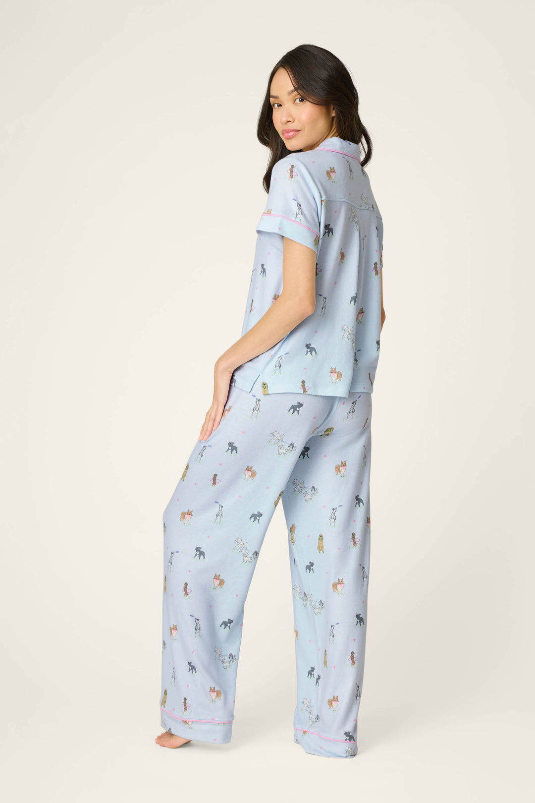 A woman stands barefoot, turned slightly to the side, wearing the P.J. Salvage Bark Park Fetch & Frolic 3-Piece PJ Set—light blue dog-themed sleepwear—against a plain off-white background. Ideal for playful women’s loungewear.