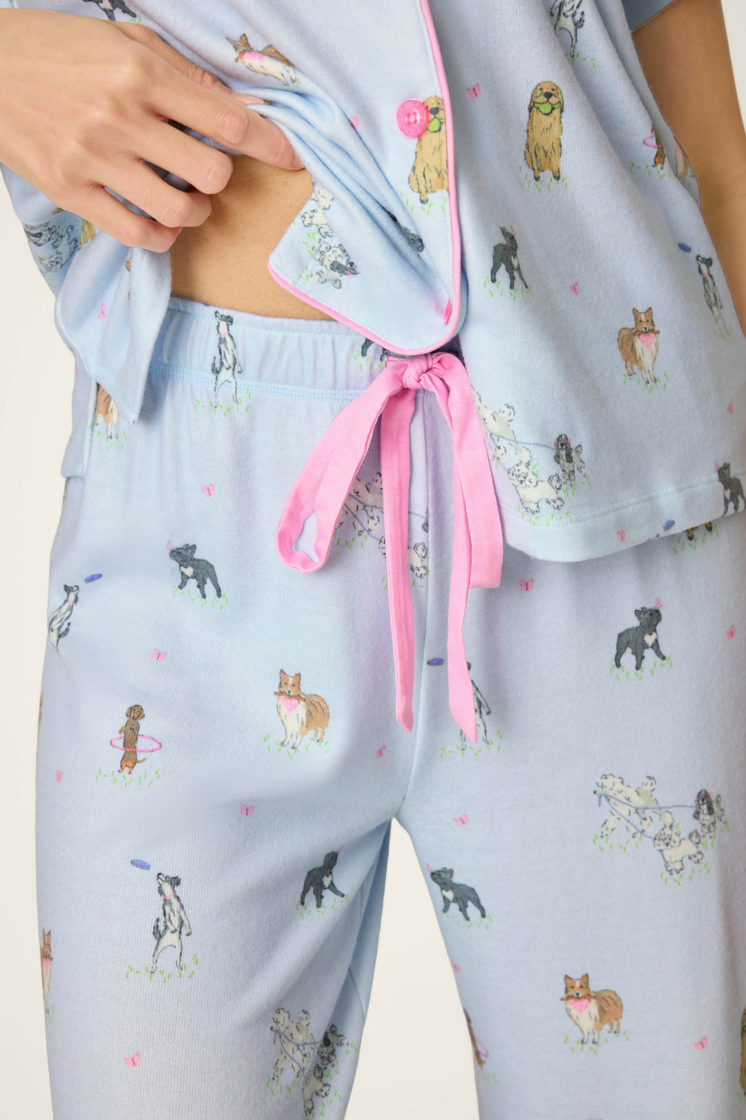 A woman wears the P.J. Salvage Bark Park Fetch & Frolic 3-Piece PJ Set, a light blue sleepwear set with pink trim, playful dog print featuring various breeds and hearts, plus a pink waist tie visible as the top is lifted. Perfect for dog lovers!.