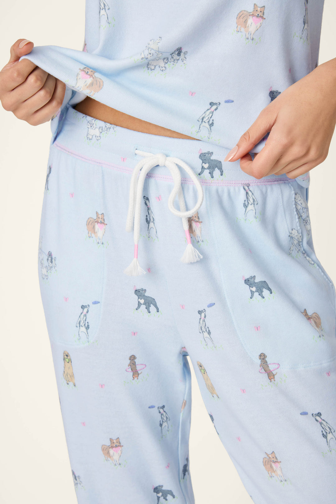 A person wears P.J. Salvage Bark Park Fetch & Frolic Banded Pants—light blue with pup print and white drawstring—holding up the top to show the waistband. Only torso and upper legs are visible, highlighting these cozy lounge pants for relaxing.