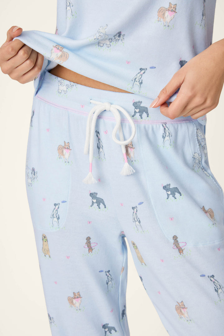 A person wears P.J. Salvage Bark Park Fetch & Frolic Banded Pants—light blue with pup print and white drawstring—holding up the top to show the waistband. Only torso and upper legs are visible, highlighting these cozy lounge pants for relaxing.
