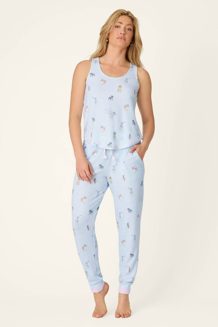 A woman stands barefoot in P.J. Salvage's Bark Park Fetch & Frolic Banded Pants—a light blue pajama jogger with playful animal prints. She has one hand in her pocket and a relaxed expression.