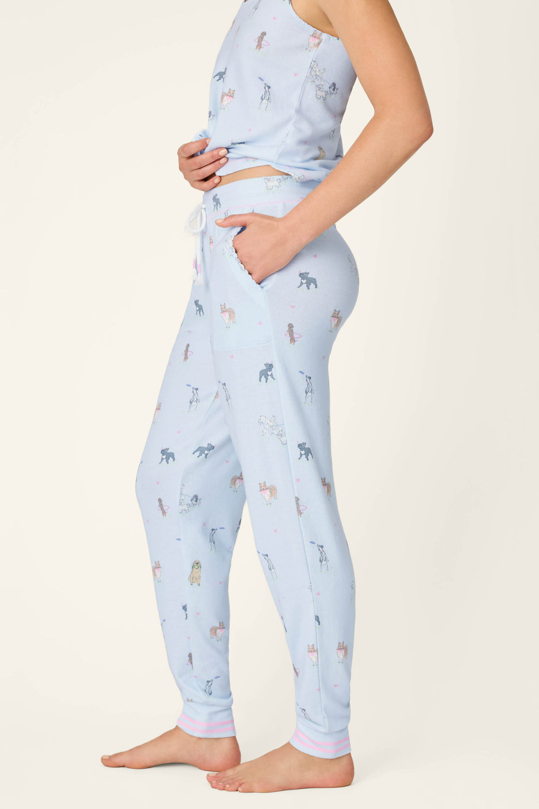A woman stands barefoot in P.J. Salvage Bark Park Fetch & Frolic Banded Pants—a light blue jersey set with a playful pup print and matching tank top. One hand’s in her pocket, the plain background highlighting the cozy pajama look.