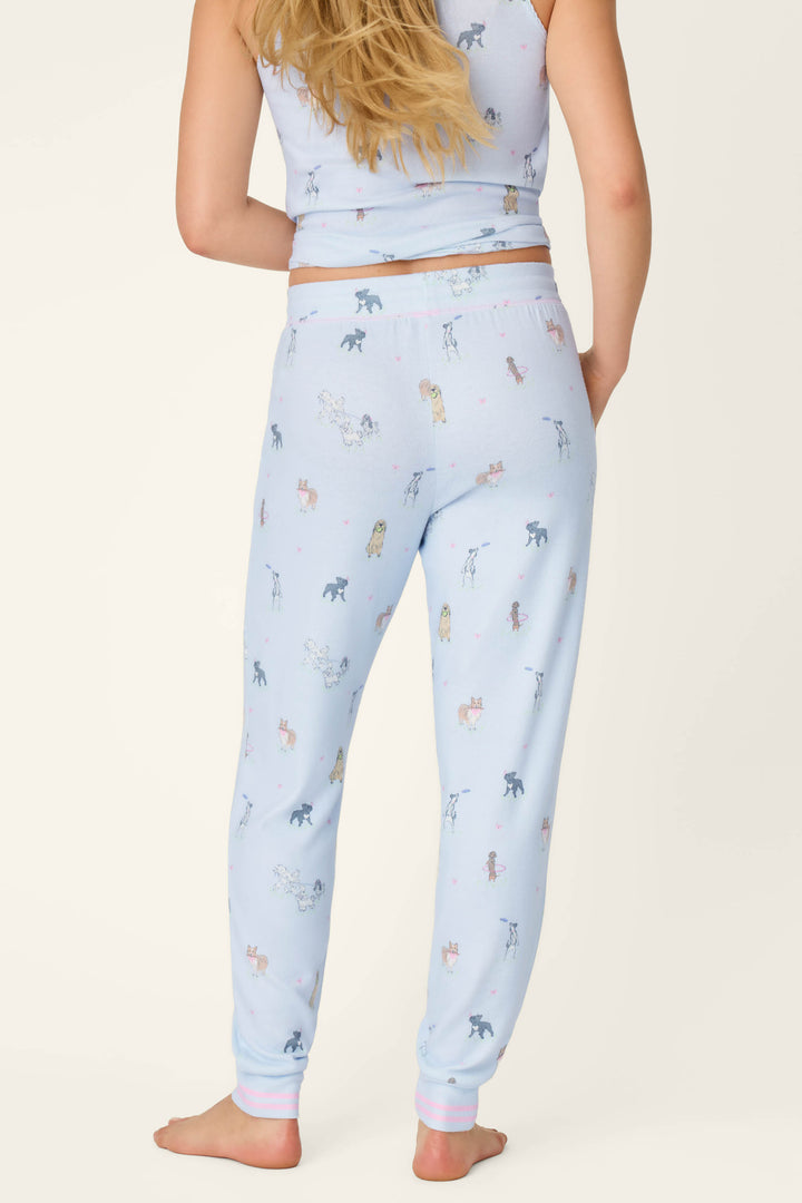 A woman with long blonde hair stands barefoot against a plain, light background, wearing P.J. Salvage’s Bark Park Fetch & Frolic Banded Pants and a matching top with a playful dog print.