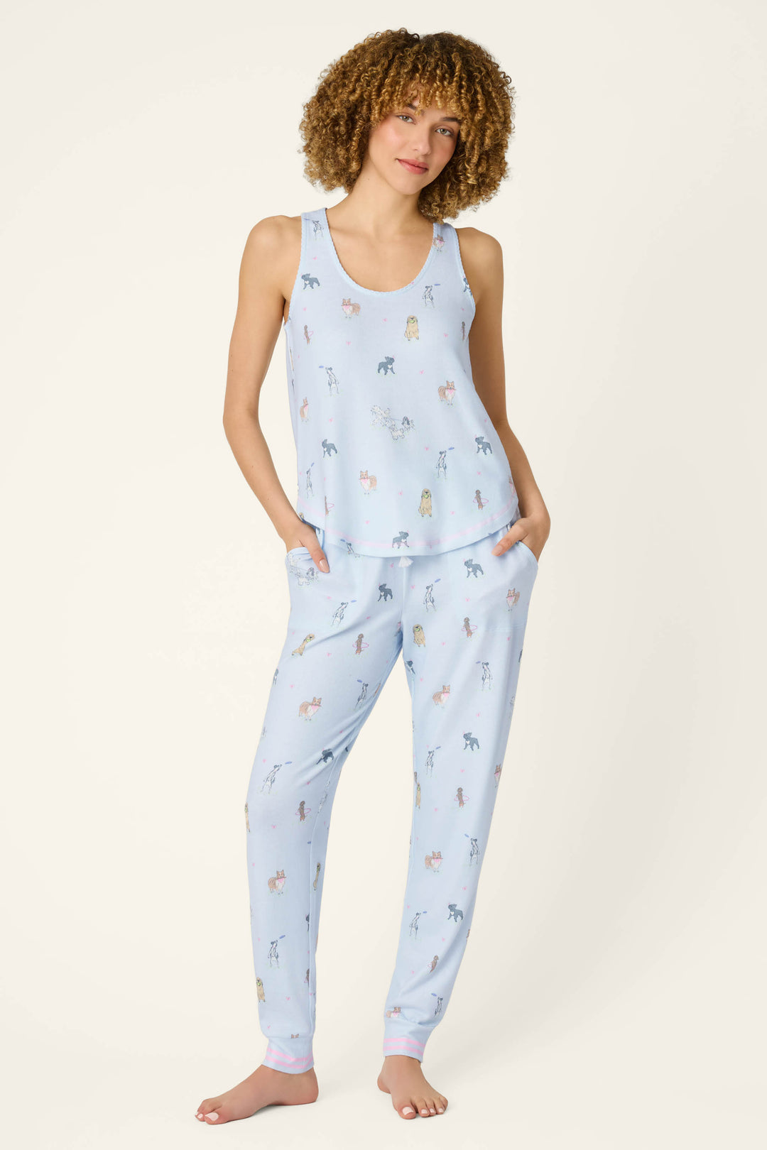 A curly-haired person wears the P.J. Salvage Bark Park Fetch & Frolic Long PJ Set—peachy-soft light blue jogger pants and a tank top with playful animal prints—standing barefoot, hands in pockets, against a plain light background.