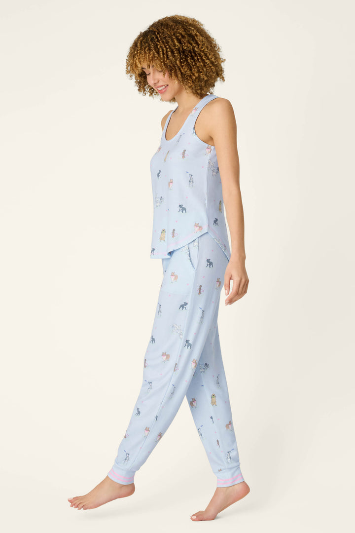 A woman smiles while walking barefoot, wearing P.J. Salvage’s Bark Park Fetch & Frolic Long PJ Set—a light blue, peachy-soft jersey pajama with a tank top and jogger pants featuring a playful dog print on a plain background.