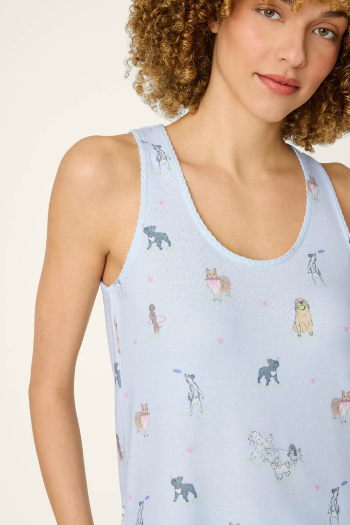 A woman with curly hair smiles softly, wearing the P.J. Salvage Bark Park Fetch & Frolic Long PJ Set top—a light blue sleeveless piece adorned with colorful dog breeds and tiny pink bows. Only her upper body is visible.