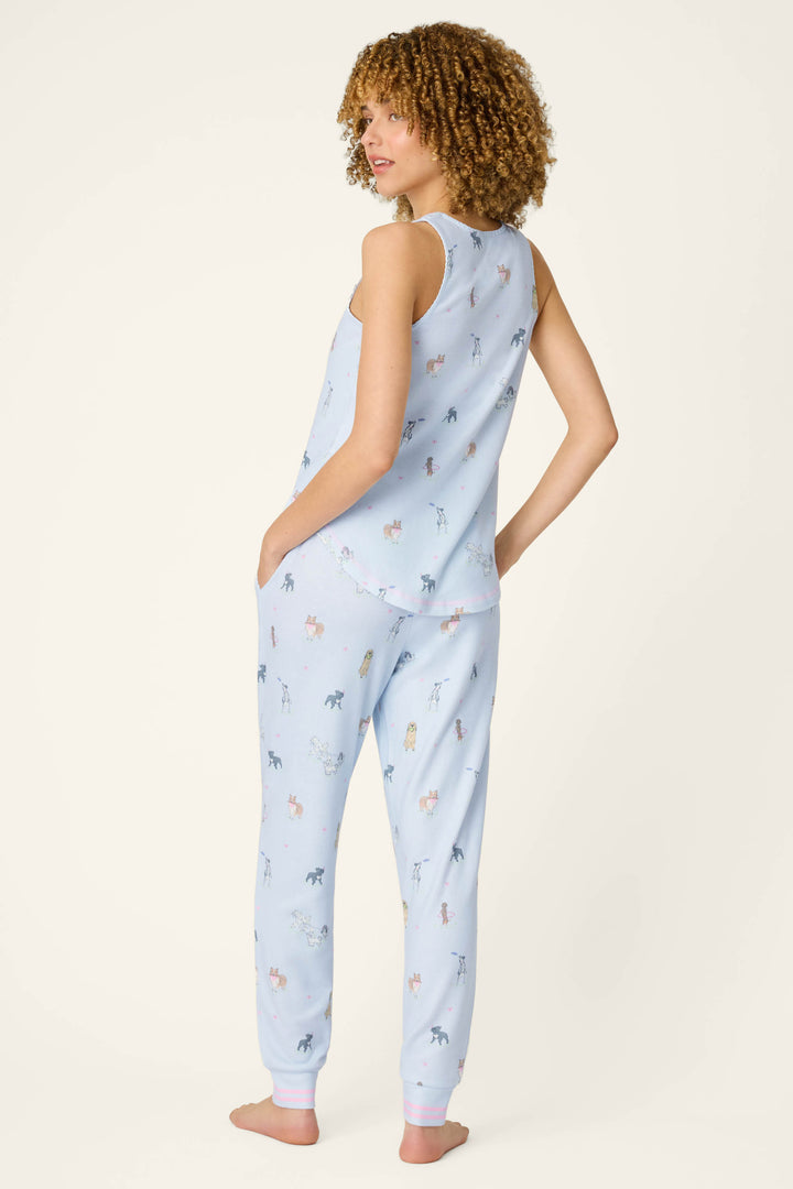 A woman with curly hair stands barefoot, facing slightly away from the camera, wearing the P.J. Salvage Bark Park Fetch & Frolic Long PJ Set—light blue top with jogger-style pants in peachy-soft jersey. The background is plain and light-colored.