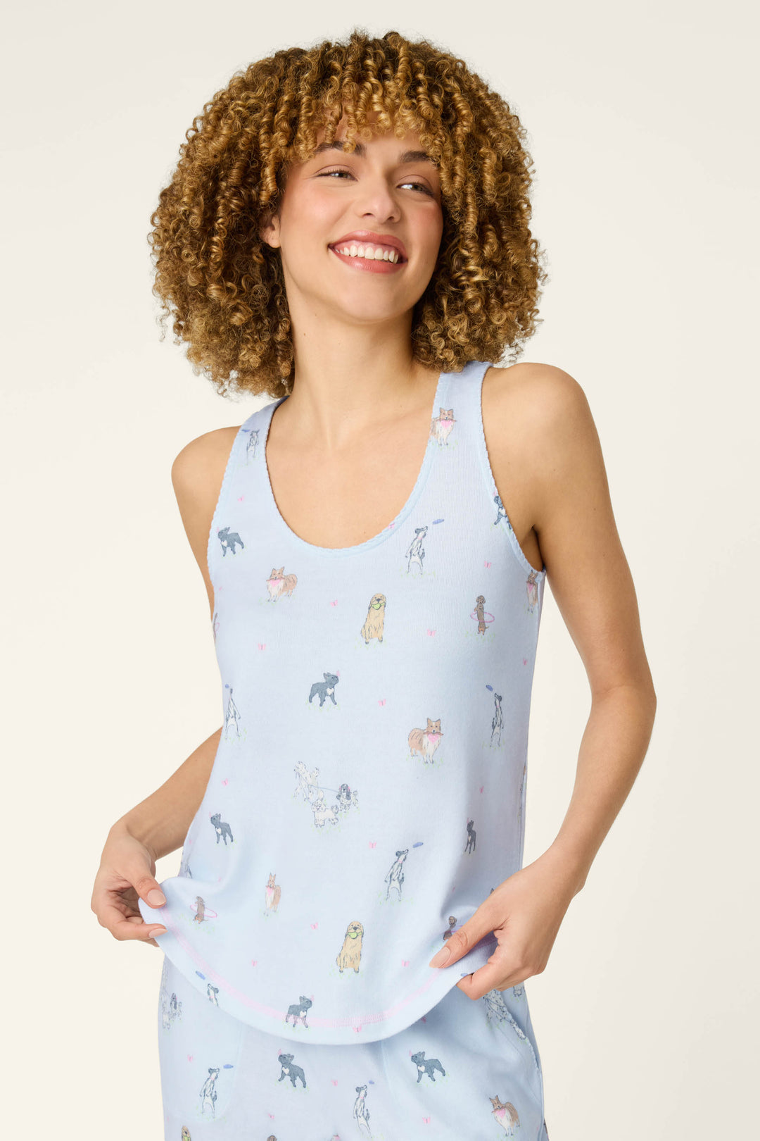A smiling woman with curly hair wears the P.J. Salvage Bark Park Fetch & Frolic Tank Top, a cozy women’s loungewear piece featuring a light blue dog print and tiny pink hearts as she holds the hem of her shirt.