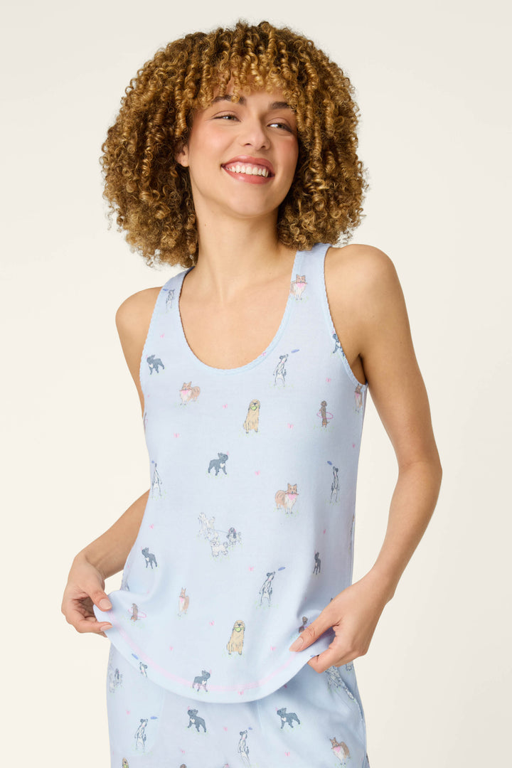 A smiling woman with curly hair wears the P.J. Salvage Bark Park Fetch & Frolic Tank Top, a cozy women’s loungewear piece featuring a light blue dog print and tiny pink hearts as she holds the hem of her shirt.