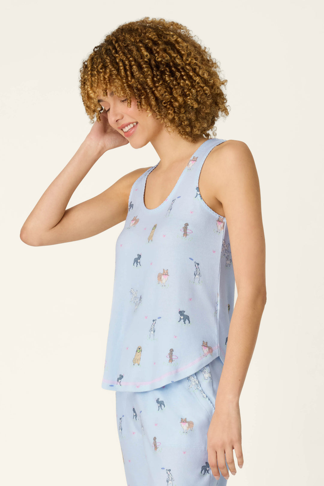 A young woman with curly hair smiles in the P.J. Salvage Bark Park Fetch & Frolic Tank Top, a light blue women's loungewear piece with a playful dog print, paired with matching bottoms and set against a plain background.