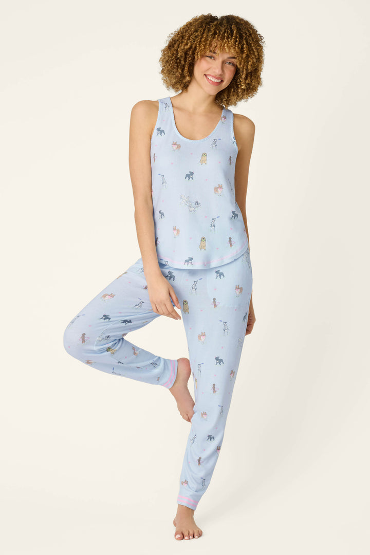 A curly-haired woman smiles while wearing P.J. Salvage's Bark Park Fetch & Frolic Tank Top—a light blue women’s sleep tank with a multicolored animal print—paired with matching pajama pants, standing with one foot against her knee.