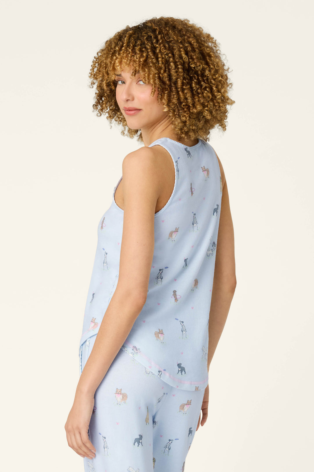 A woman with curly hair models the P.J. Salvage Bark Park Fetch & Frolic Tank Top, a light blue sleep tank with a colorful dog print, as she stands against a plain background and looks over her shoulder.