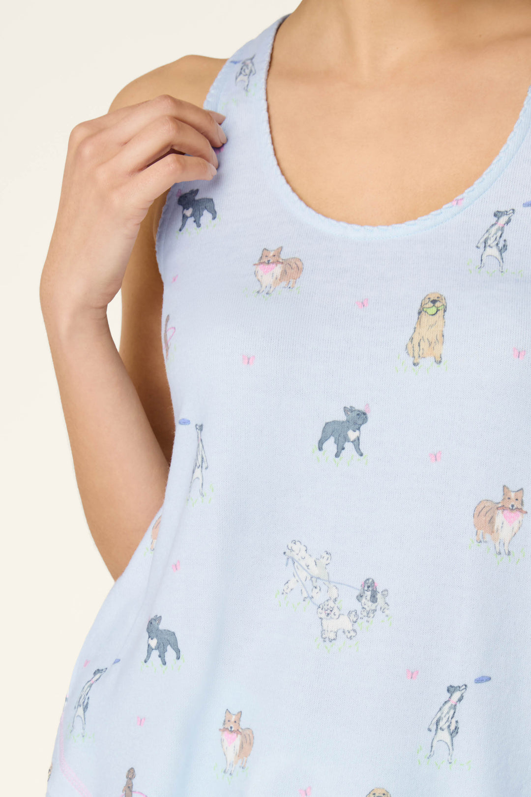 A person wears the P.J. Salvage Bark Park Fetch & Frolic Tank Top, a light blue women's loungewear sleep tank featuring a playful dog print. Their hand gently touches the strap near their shoulder.