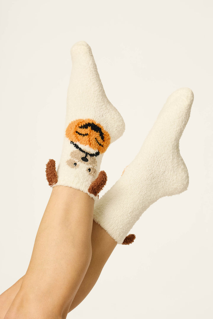Two crossed legs wearing fuzzy white "Boyfriend Boxer Trick-or-Treating Dog Socks" by P.J. Salvage, each featuring an adorable dog face with orange pumpkin accents on the front and small brown ear and tail details. The background is plain and light-colored.