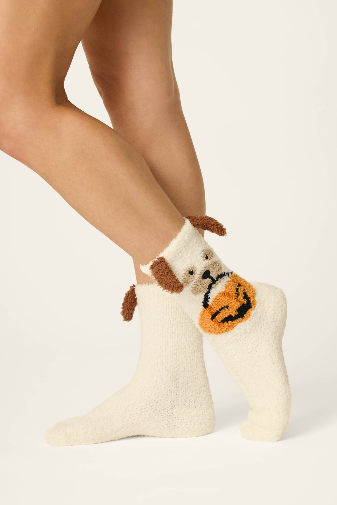 A person stands on tiptoe against a plain off-white background, wearing P.J. Salvage Boyfriend Boxer Trick-or-Treating Dog Socks—cozy cream-colored socks featuring a dog face with floppy brown ears and an orange pumpkin decoration on the side.