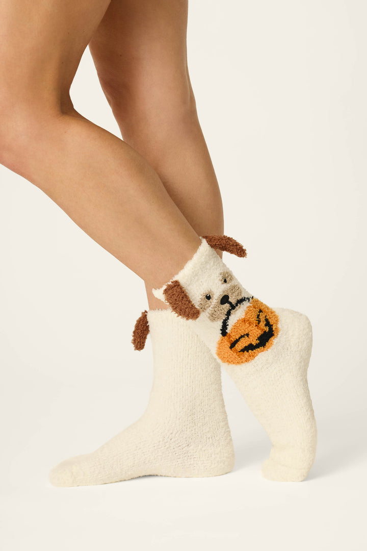 A person stands on tiptoe against a plain off-white background, wearing P.J. Salvage Boyfriend Boxer Trick-or-Treating Dog Socks—cozy cream-colored socks featuring a dog face with floppy brown ears and an orange pumpkin decoration on the side.