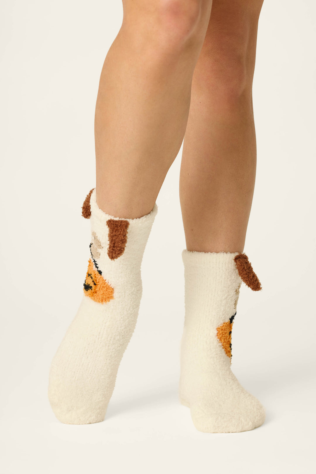 A person wearing P.J. Salvage Boyfriend Boxer Trick-or-Treating Dog Socks, featuring fuzzy white fabric with brown floppy dog ears and an orange dog face design, stands on a light background.