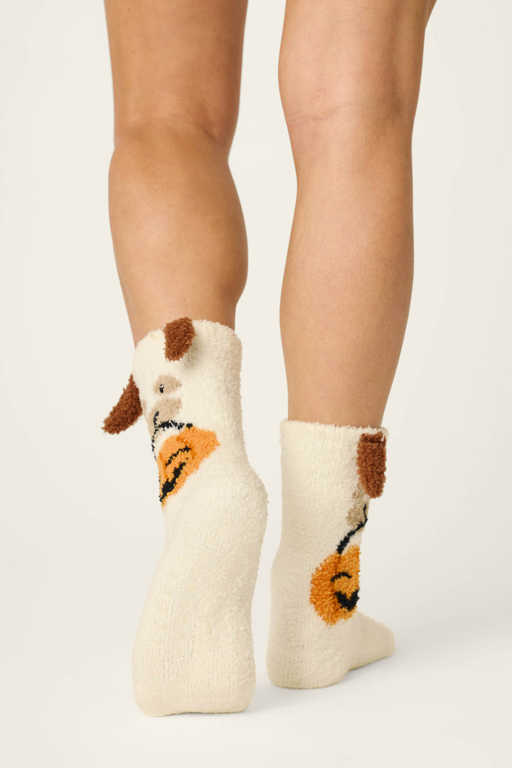 A person stands on tiptoes wearing P.J. Salvage Boyfriend Boxer Trick-or-Treating Dog Socks featuring a white fuzzy texture, brown ears, and a stitched orange nose design; the background is plain and light-colored.