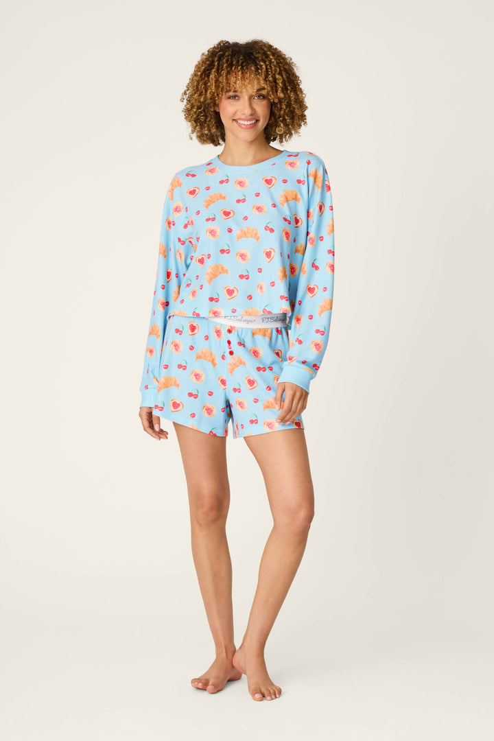 A person with curly hair, barefoot and smiling, stands against a plain light background wearing the P.J. Salvage Boyfriend Boxer Pastry Print Long Sleeve & Shorts PJ Set in light blue, featuring a colorful food print.