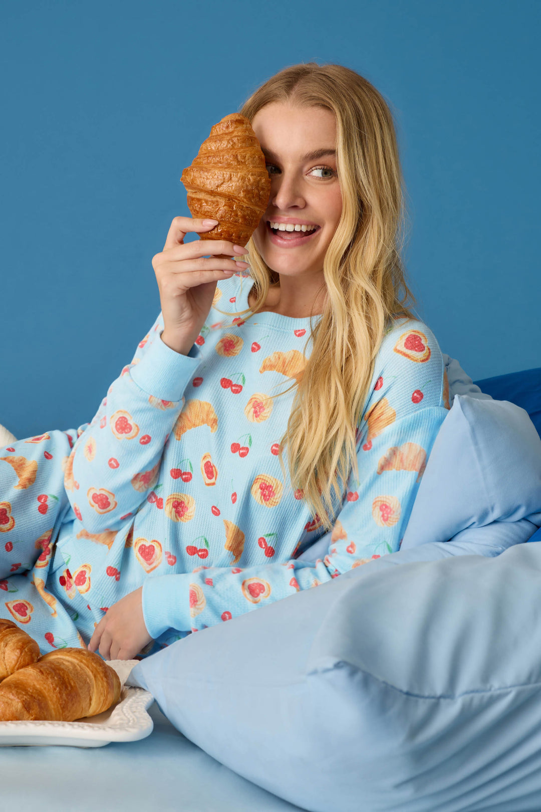 Wearing the P.J. Salvage Boyfriend Boxer Pastry Print Long Sleeve & Shorts PJ Set, a woman sits on a bed with blue sheets and background, smiling as she holds a croissant over one eye. A plate of croissants rests by her side.