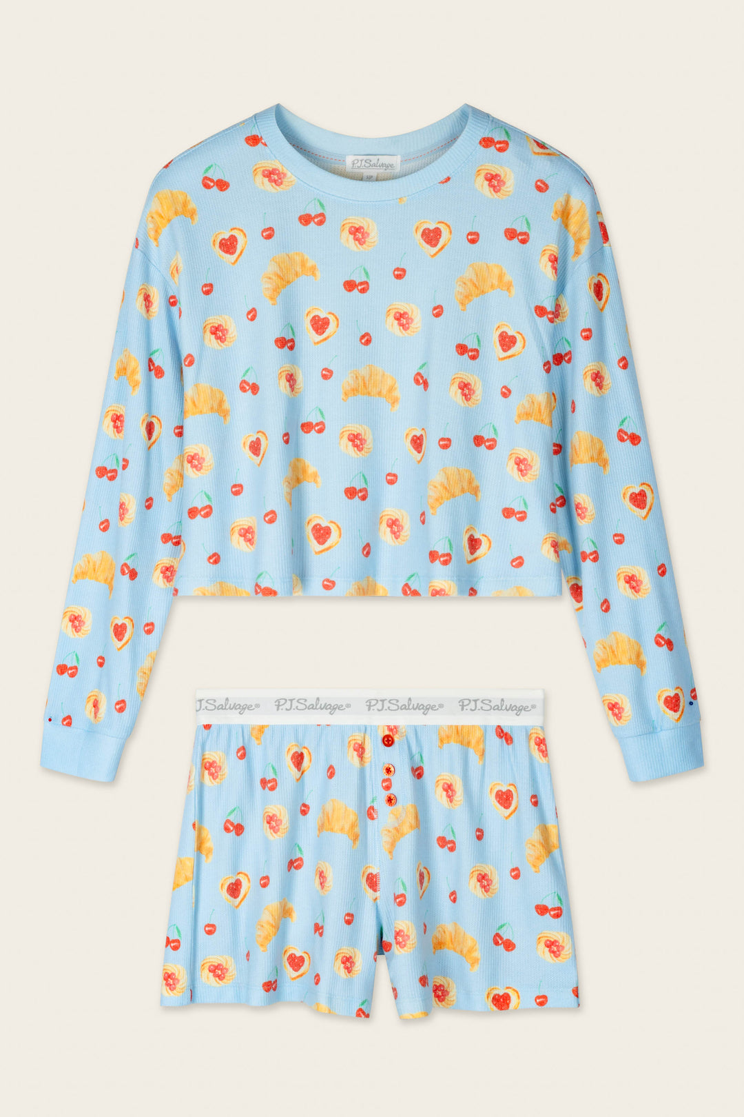 The P.J. Salvage Boyfriend Boxer Pastry Print Long Sleeve & Shorts PJ Set features a light blue long-sleeve top and shorts decorated with playful prints of croissants, hearts, and cherries. The shorts include a white elastic waistband printed with "PJ Salvage." The set is laid flat on a beige background.